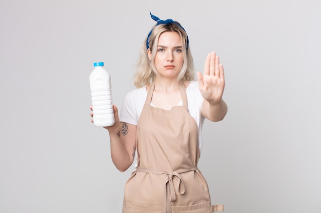 Młoda ładna kobieta albinos wygląda poważnie pokazując otwartą dłoń, robiąc gest zatrzymania z butelką mleka