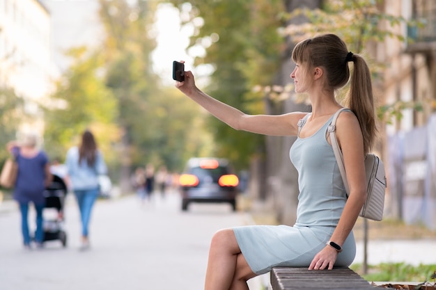 Młoda ładna kobieta biorąc selfie z telefonu komórkowego w ciepły letni dzień, siedząc na ławce ulicy miasta na zewnątrz.
