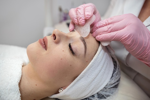 Młoda ładna kobieta otrzymuje masaż twarzy z zabiegiem guasha w klinice kosmetycznej