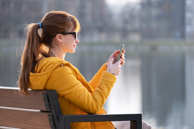 Młoda ładna kobieta siedzi na ławce w parku, przeglądając jej smartfona na zewnątrz w ciepły jesienny dzień.
