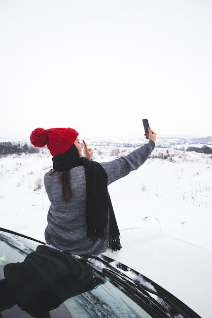 Młoda ładna kobieta siedzi na maskę samochodu i biorąc selfie. podróż samochodem terenowym. styl życia