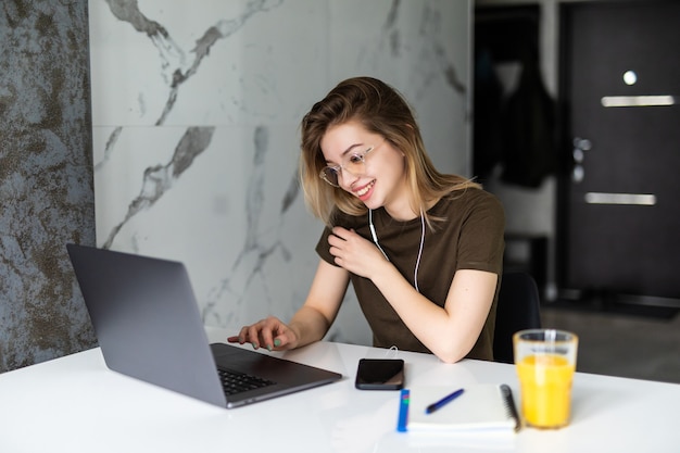 Młoda ładna kobieta siedzi przy stole w kuchni za pomocą laptopa i używać na telefonie komórkowym.