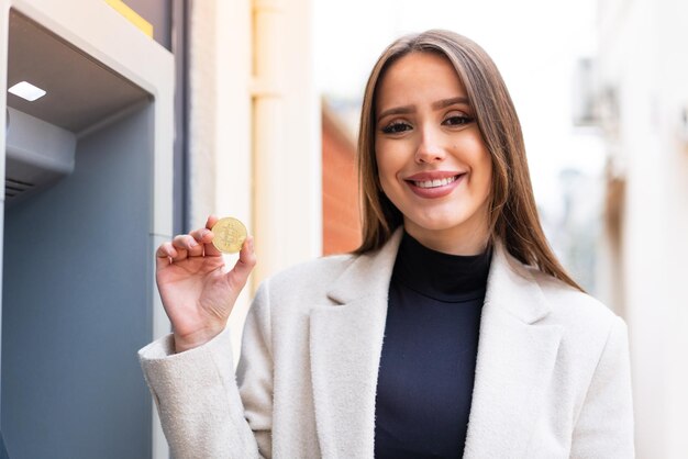 Młoda ładna kobieta trzyma Bitcoina na zewnątrz, dużo się uśmiechając