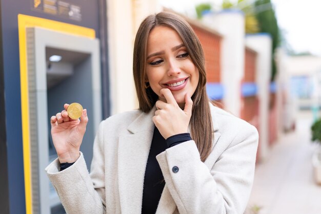 Młoda ładna kobieta trzyma Bitcoina na zewnątrz, myśląc o pomyśle i patrząc z boku