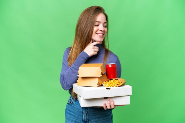 Młoda ładna kobieta trzymająca pizze i hamburgery na odizolowanym tle klucza chrominancji, patrząca z boku i uśmiechająca się