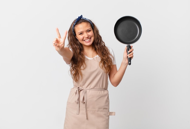 Młoda ładna kobieta uśmiecha się i wygląda na szczęśliwą, gestykuluje zwycięstwo lub koncepcję pokoju szefa kuchni i trzyma patelnię