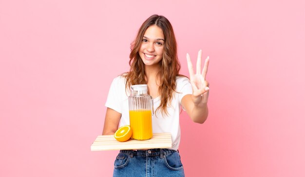 Młoda ładna kobieta uśmiechnięta i wyglądająca przyjaźnie, pokazująca numer trzy i trzymająca tacę z sokiem pomarańczowym