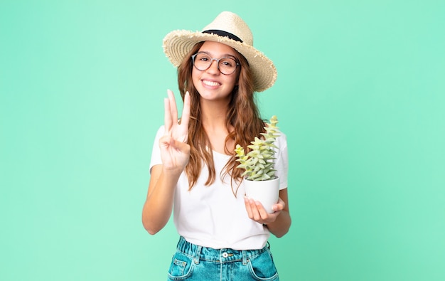 Młoda ładna kobieta uśmiechnięta i wyglądająca przyjaźnie, pokazująca numer trzy w słomkowym kapeluszu i trzymająca kaktusa