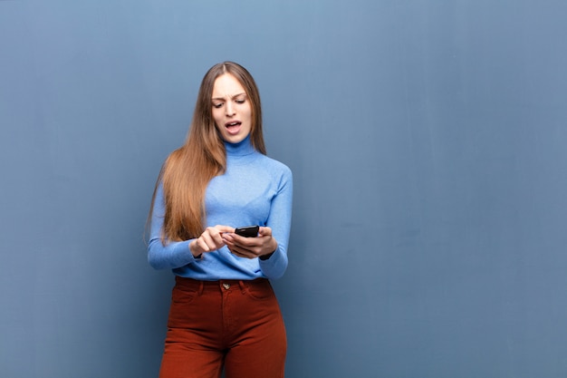 Młoda ładna kobieta używa mądrze telefon przeciw błękit ścianie z odbitkową przestrzenią