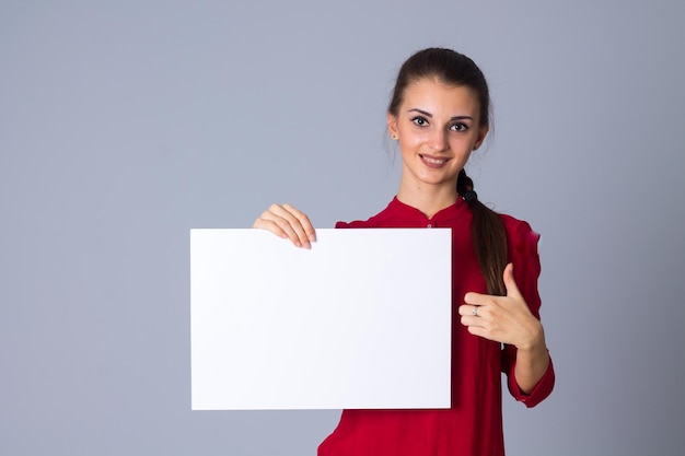 Młoda ładna kobieta w czerwonej bluzce trzyma białą kartkę papieru na szarym tle w studio