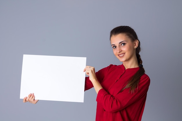 Młoda ładna kobieta w czerwonej bluzce z warkoczem, trzymająca białą kartkę papieru na szarym tle w studio