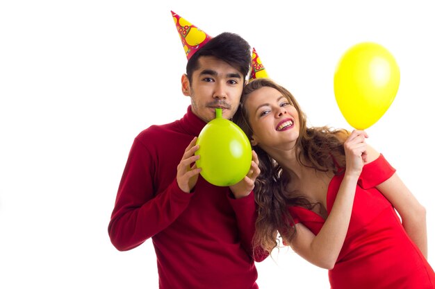 Młoda ładna kobieta w czerwonej sukience i młody przystojny mężczyzna w kolorowych kapeluszach dmuchający balony
