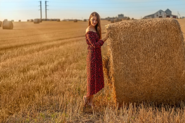 Młoda ładna kobieta w czerwonej sukience z bliska na tle pola z stogi w czasie zachodu słońca.