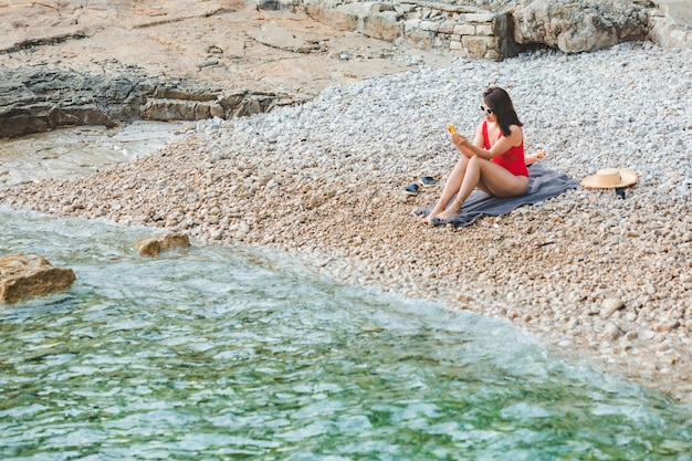 Młoda ładna kobieta w czerwonym stroju kąpielowym siedzi na kamienistej plaży z telefonem morze letnie wakacje