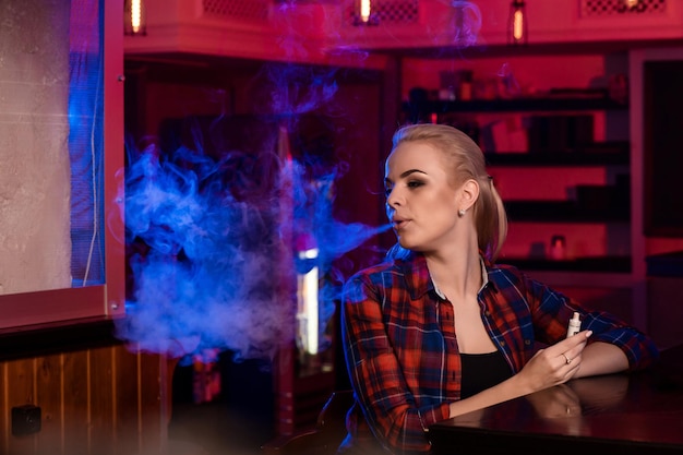 Młoda ładna kobieta w koszuli w klatce pali elektronicznego papierosa w barze vape