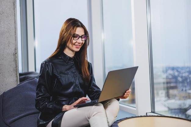 Młoda ładna kobieta w okularach pracuje na laptopie, siedząc w nowoczesnym obszarze roboczym Praca zdalna jako freelancer