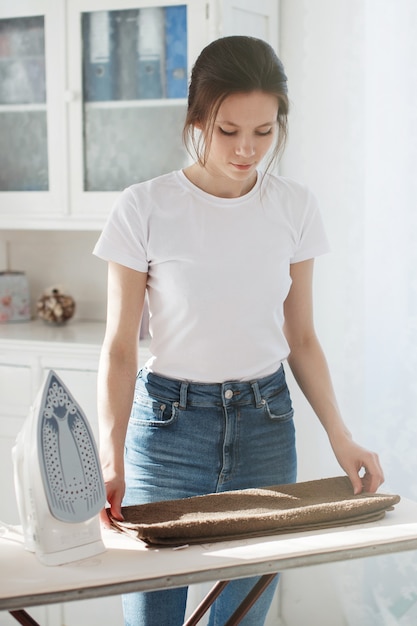 Zdjęcie młoda ładna kobieta w t-shircie i dżinsach gładziła bieliznę i układała ubrania w stosy. w pobliżu żelazko, za nim biały bufet.