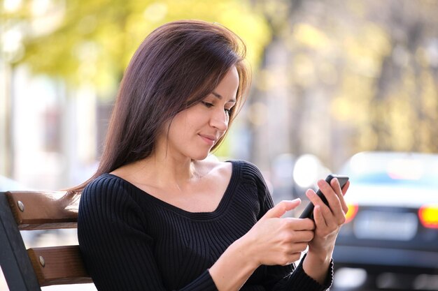 Młoda ładna Kobieta Wiadomości Na Telefon Komórkowy W Ciepły Letni Dzień, Siedząc Na Miejskiej ławce Na świeżym Powietrzu