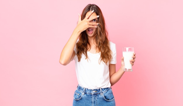 Zdjęcie młoda ładna kobieta wygląda na zszokowaną, przestraszoną lub przerażoną, zakrywa twarz dłonią i trzyma szklankę mleka