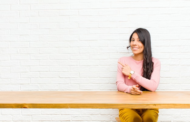 Młoda ładna kobieta z Ameryki Łacińskiej, uśmiechając się wesoło, czując się szczęśliwa i wskazując na bok i do góry, pokazując przedmiot w przestrzeni kopii przed stołem