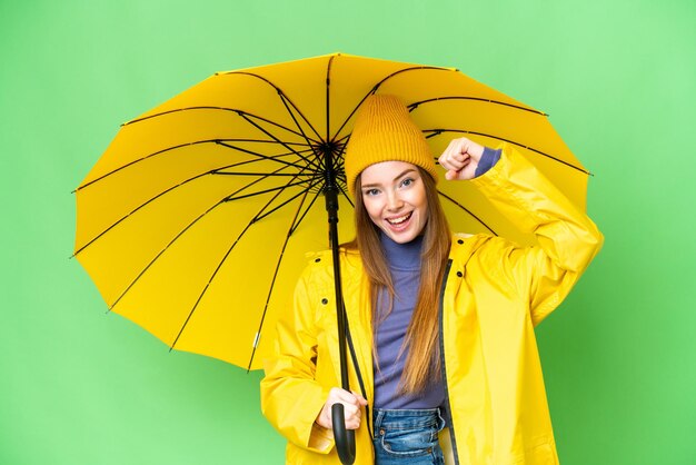 Młoda ładna kobieta z płaszczem przeciwdeszczowym i parasolem na odizolowanym tle klucza chrominancji świętuje zwycięstwo