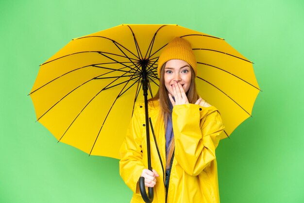 Młoda ładna kobieta z płaszczem przeciwdeszczowym i parasolem na odizolowanym tle klucza chrominancji, szczęśliwa i uśmiechnięta, zakrywająca usta dłonią