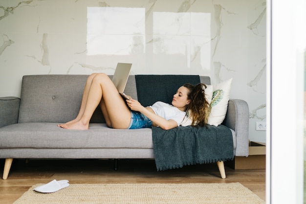 młoda ładna kobieta zakupy online na laptopie leży na kanapie, pisząc na laptopie