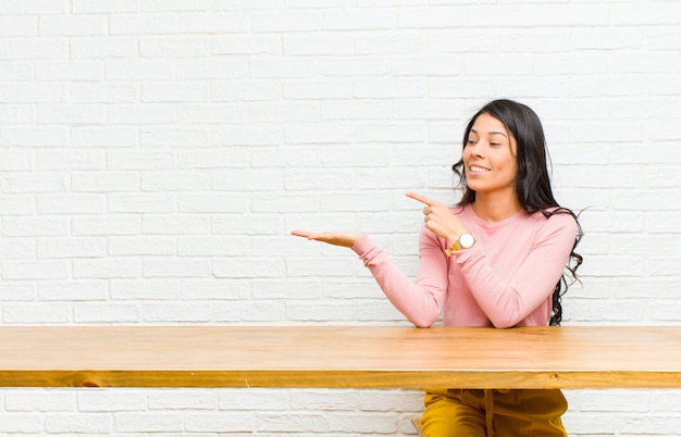 Młoda ładna łacińska kobieta ono uśmiecha się radośnie i wskazuje copyspace na palmie na stronie, pokazuje lub reklamuje przedmiot siedzi przed stołem