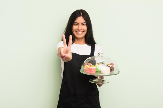 Młoda ładna latynoska kobieta uśmiecha się i wygląda przyjaźnie pokazując szefa kuchni numer dwa z koncepcją ciast!