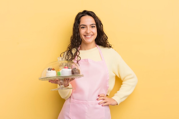 Młoda ładna latynoska kobieta uśmiecha się szczęśliwie z ręką na biodrze i pewna siebie koncepcja szefa kuchni i ciast
