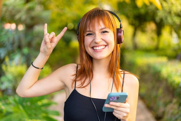 Młoda ładna rudowłosa kobieta na zewnątrz słucha muzyki za pomocą telefonu komórkowego wykonującego rockowy gest