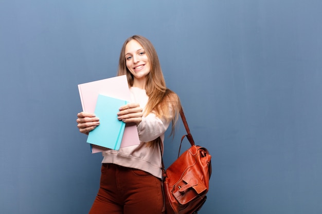 Młoda ładna studencka kobieta z książkami i torbą przeciw błękit ścianie z odbitkową przestrzenią