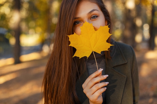 Młoda ładna stylowa kobieta o niebieskich oczach w modnym płaszczu, trzymając żółty jesienny liść przy twarzy podczas spaceru po parku. Szczegół upadek portret szczęśliwa dziewczyna