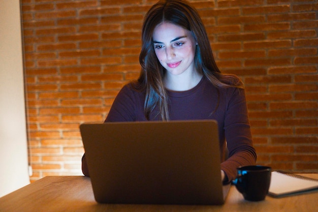 Młoda, ładnie uśmiechnięta kobieta używająca laptopa w domu w nocy Szczęśliwa dziewczyna patrząca na komputer Wesołe ludzie siedzący przy biurku w salonie Koncepcja przedsiębiorczej pracy zdalnej i ciężko pracującej osoby