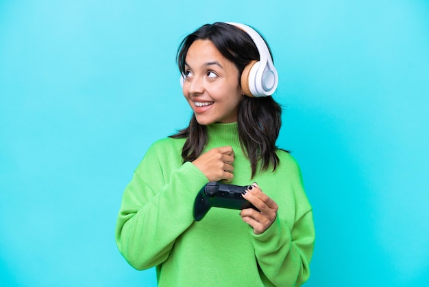 Młoda latynoska kobieta bawi się kontrolerem gier wideo odizolowanym na niebieskim tle, patrząc w górę, uśmiechając się