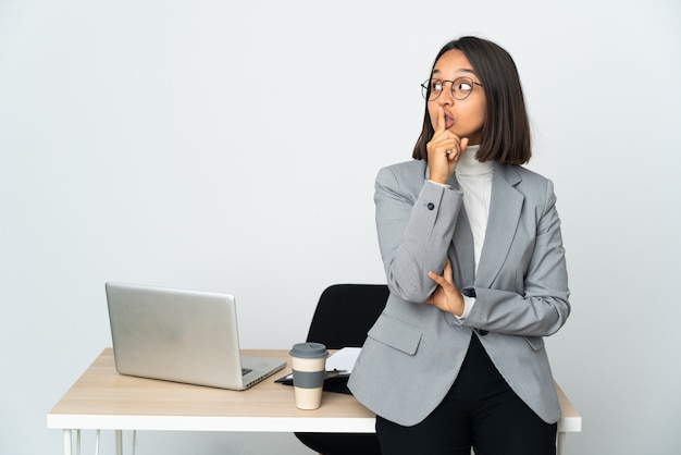 Młoda latynoska kobieta biznesu pracująca w biurze na białym tle pokazująca znak ciszy gest wkładania palca do ust