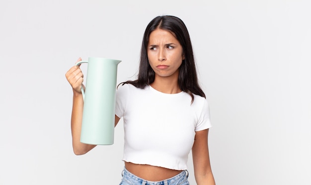 Młoda latynoska kobieta czuje się smutna, zdenerwowana lub zła, patrzy w bok i trzyma termos z kawą