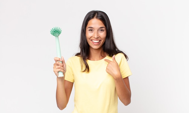 Młoda latynoska kobieta czuje się szczęśliwa i wskazuje na siebie podekscytowaną i trzymającą szczoteczkę do czyszczenia naczyń