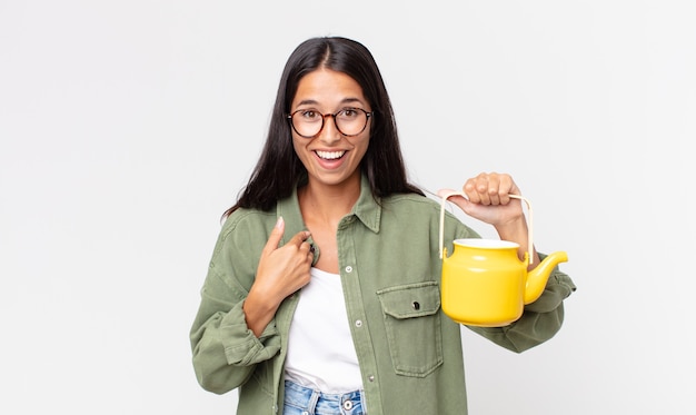 Młoda latynoska kobieta czuje się szczęśliwa i wskazuje na siebie z podekscytowanym i trzymającym dzbanek do herbaty