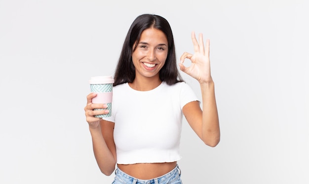 Młoda latynoska kobieta czuje się szczęśliwa, pokazując aprobatę dobrym gestem i trzymająca pojemnik na kawę na wynos
