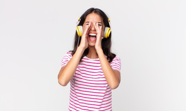 Młoda latynoska kobieta czuje się szczęśliwa, wydając wielki okrzyk z rękami przy ustach, słuchając muzyki przez słuchawki