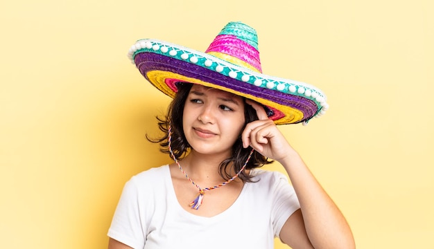 Zdjęcie młoda latynoska kobieta czuje się zdezorientowana i zdezorientowana, drapiąc się po głowie. koncepcja meksykańskiego kapelusza