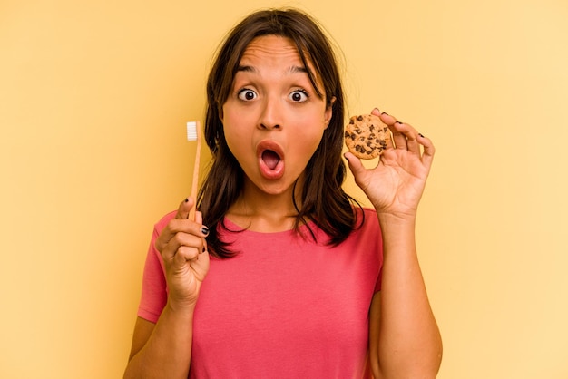 Młoda latynoska kobieta myjąca zęby po zjedzeniu ciasteczek na żółtym tle
