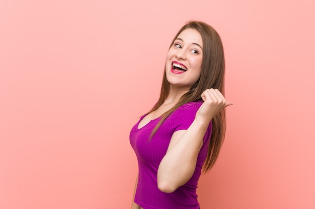 Młoda latynoska kobieta przy różowej ścianie wskazuje kciukiem palec, śmiejąc się i beztrosko.