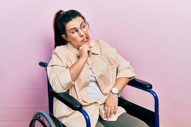 Młoda latynoska kobieta siedząca na wózku inwalidzkim, myśląca zmartwiona pytaniem i zdenerwowana ręką na brodzie