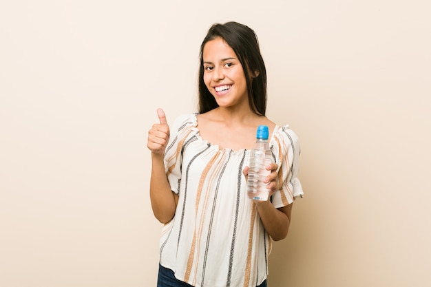 Młoda latynoska kobieta trzyma butelkę woda uśmiecha się kciuk up i podnosi