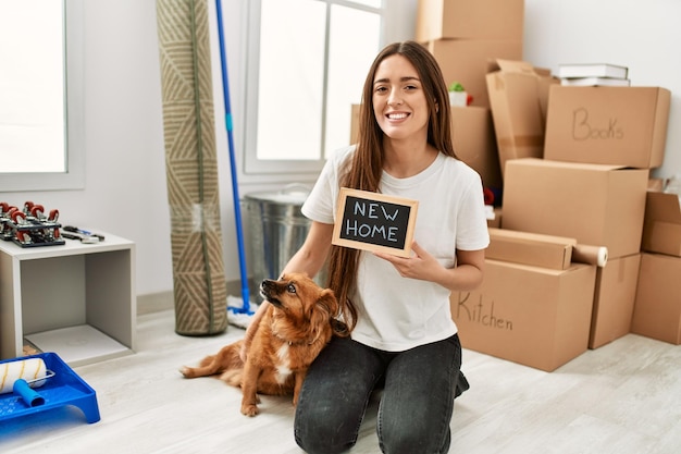 Młoda latynoska kobieta trzyma nową tablicę domową siedzącą na podłodze z psem w nowym domu