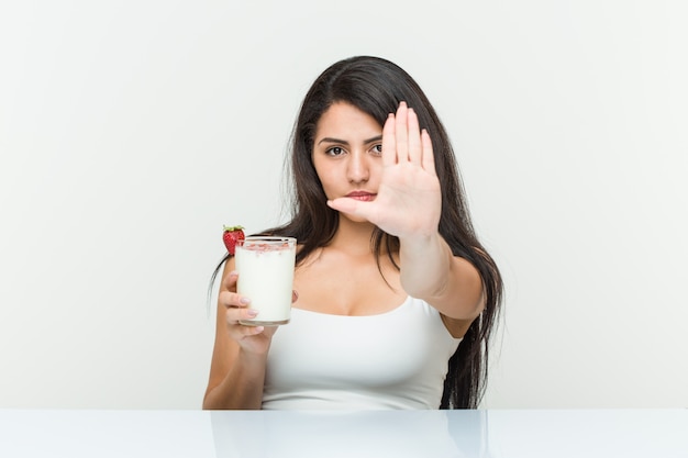 Młoda latynoska kobieta trzyma smoothie Młoda latynoska kobieta trzyma awokado toast stojący z wyciągniętą ręką pokazującą znak stopu, co zapobiega. <Mixto>