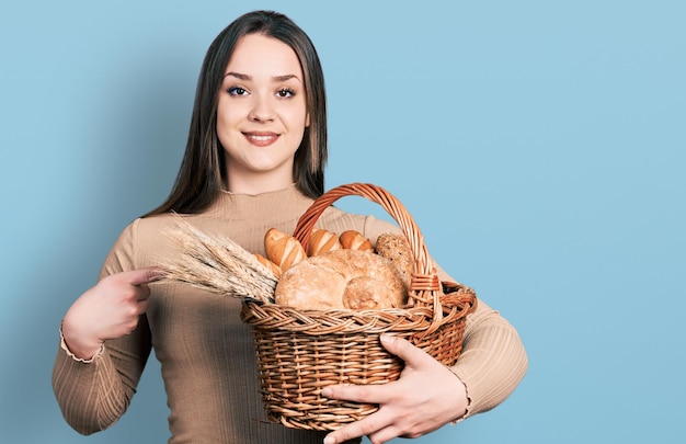 Młoda latynoska kobieta trzyma wiklinowego kosza z chlebem uśmiechniętym szczęśliwym wskazującym ręką i palcem