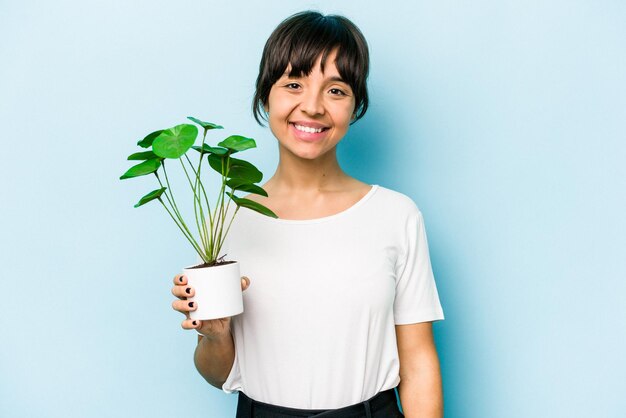 Młoda latynoska kobieta trzymająca roślinę odizolowaną na niebieskim tle szczęśliwa uśmiechnięta i wesoła